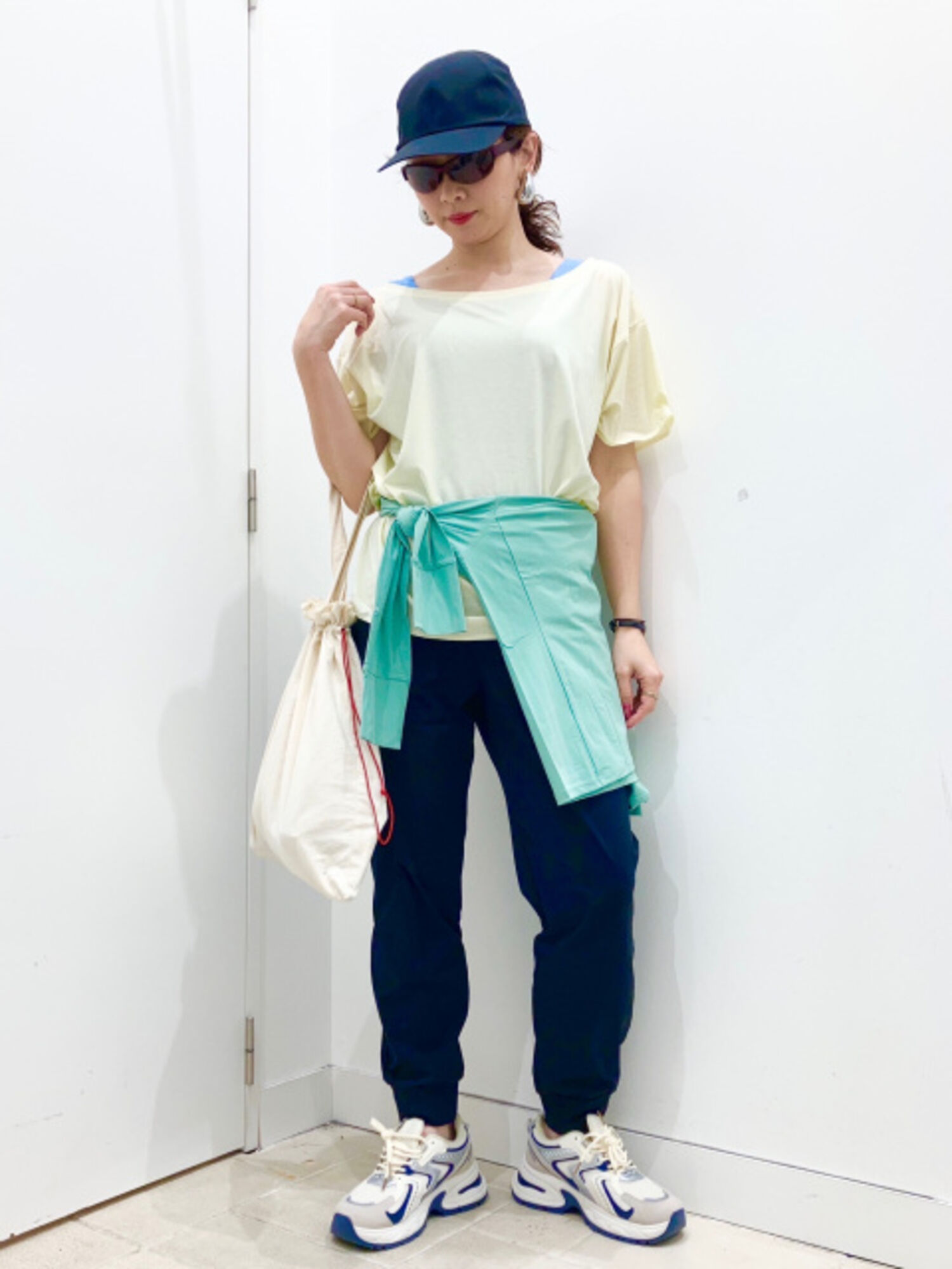 ネイビーのキャップとサングラスを着用し、レモン色のTシャツの腰にミントグリーンのパーカーを巻いた、スポーティな女性の全身コーディネート画像。